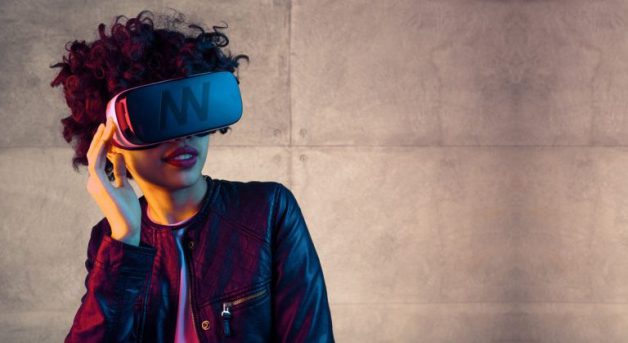 Trendy jonge vrouw in leren jack die met een grote VR-bril poseert voor een betonnen muur